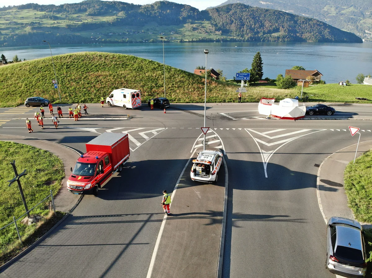 Eine Kreuzung in ländlicher Umgebung zeigt Einsatzkräfte und Fahrzeuge der Feuerwehr und Polizei nach einem Unfall. Ein See ist im Hintergrund sichtbar.