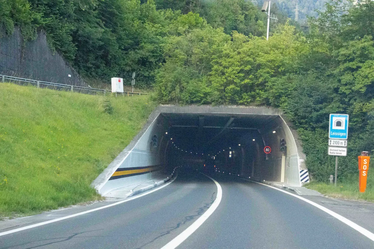 Ein Straßenabschnitt mit der Einfahrt in einen beleuchteten Tunnel. Rechts sind ein Schild mit Tunnelnamen und eine SOS-Säule sichtbar.