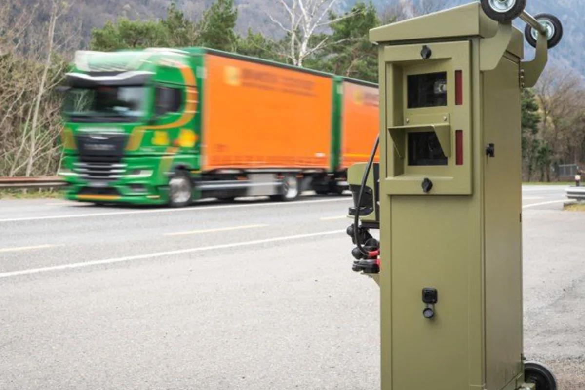 Ein mobiler Blitzer steht am Straßenrand. Ein grüner Lkw fährt mit hoher Geschwindigkeit auf der Straße vorbei. Im Hintergrund sind Bäume sichtbar.