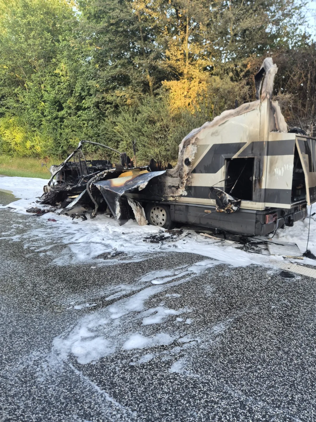 Ein stark beschädigter und ausgebrannter Wohnwagen steht auf einer Straße, umgeben von Löschschaum auf dem Asphalt.