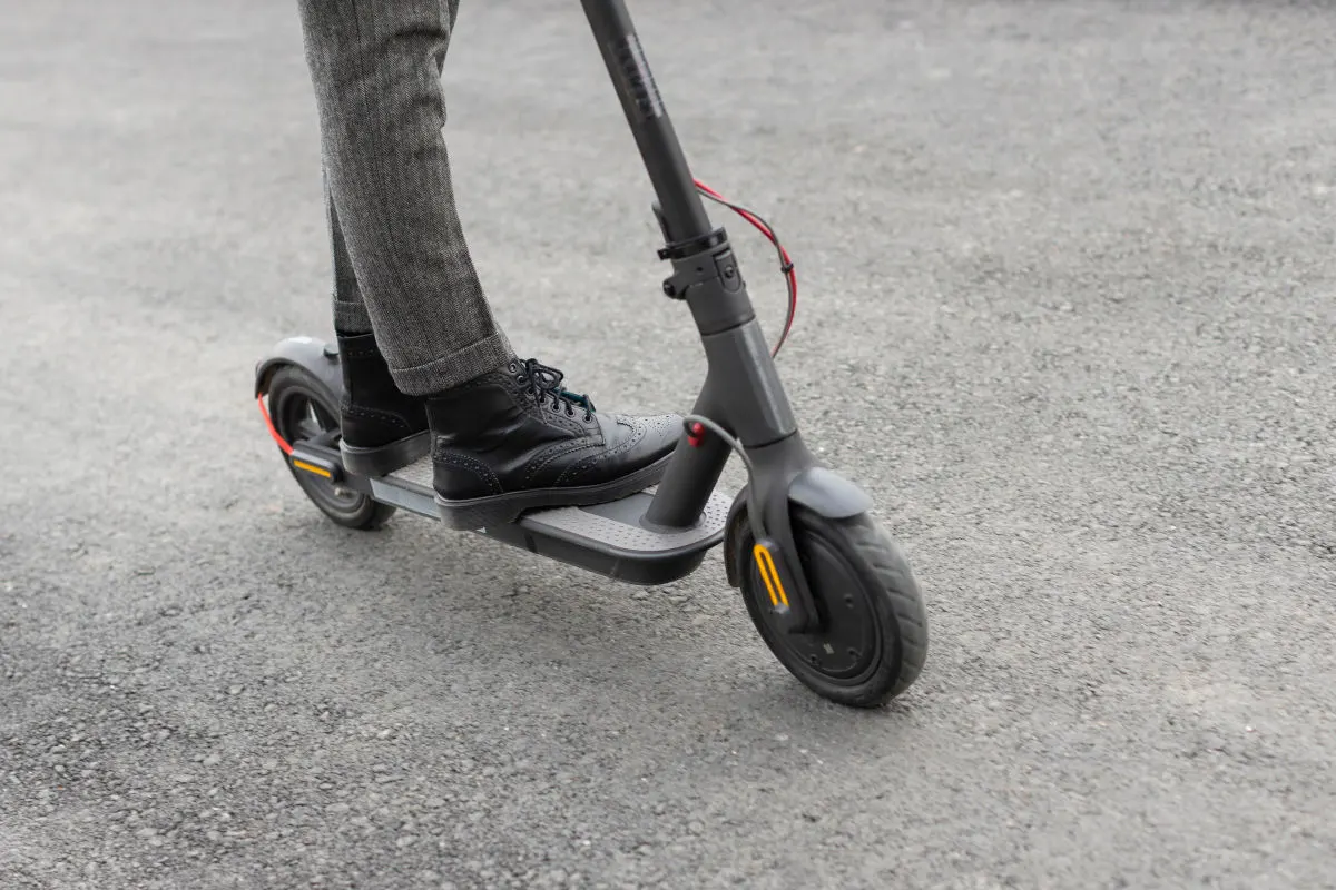 Eine Person mit grauer Hose und schwarzen Schuhen fährt auf einem Elektroroller über grauen Asphalt. Der Fokus liegt auf dem Roller und den Beinen der Person.