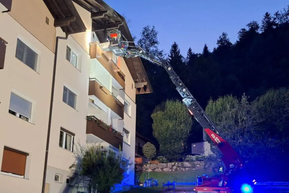 Eine Feuerwehrdrehleiter ist an einem Wohngebäude ausgefahren, während Einsatzkräfte in der Nacht arbeiten. Das Gebäude hat mehrere Stockwerke und es ist ein Wald im Hintergrund.