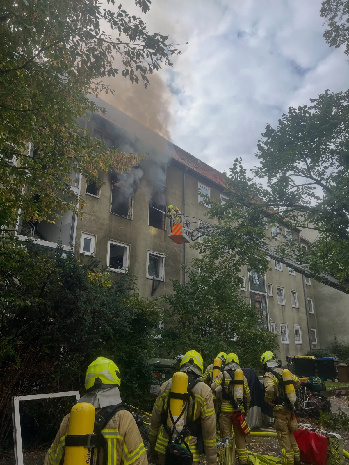 Mehrere Feuerwehrleute in voller Schutzkleidung stehen vor einem Wohngebäude, aus dessen oberen Fenstern Rauch austritt. Eine Drehleiter ist im Einsatz.