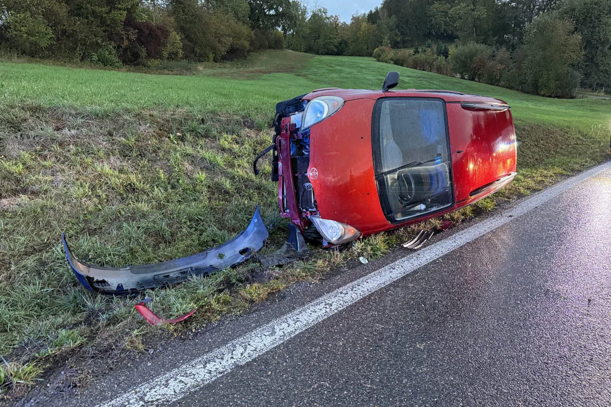 Ein rotes Auto liegt seitlich im Gras neben einer nassen Straße. Der vordere Stoßfänger ist abgerissen und im Gras zu sehen.