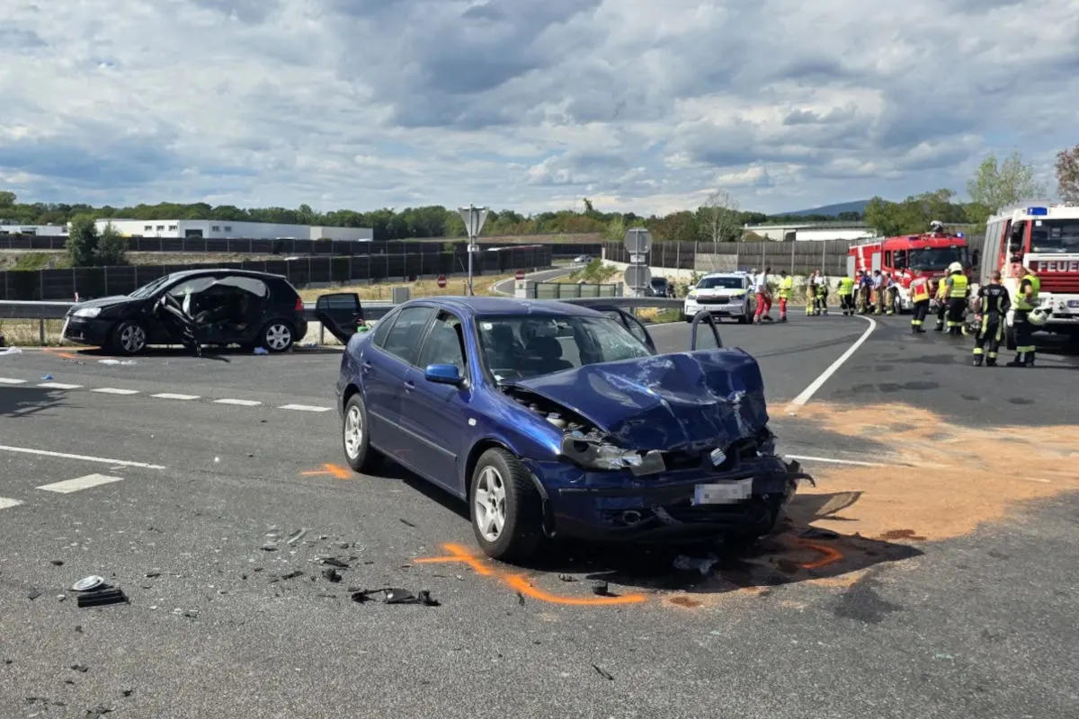 Ein schwer beschädigtes blaues Auto steht im Vordergrund auf einer Straße. Im Hintergrund sind Einsatzfahrzeuge und Feuerwehrleute zu sehen. Der Unfallort zeigt verstreute Fahrzeugteile auf dem Asphalt.