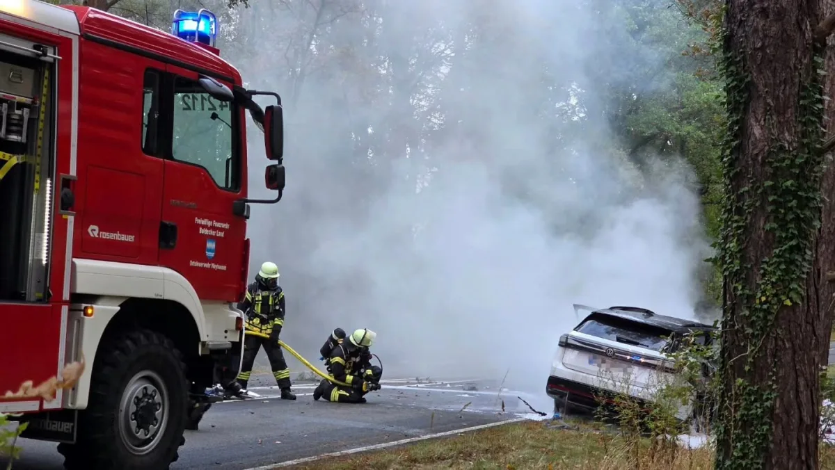 Ein Feuerwehrfahrzeug und Feuerwehrleute löschen ein stark rauchendes Auto auf einer Straße in einer bewaldeten Gegend.