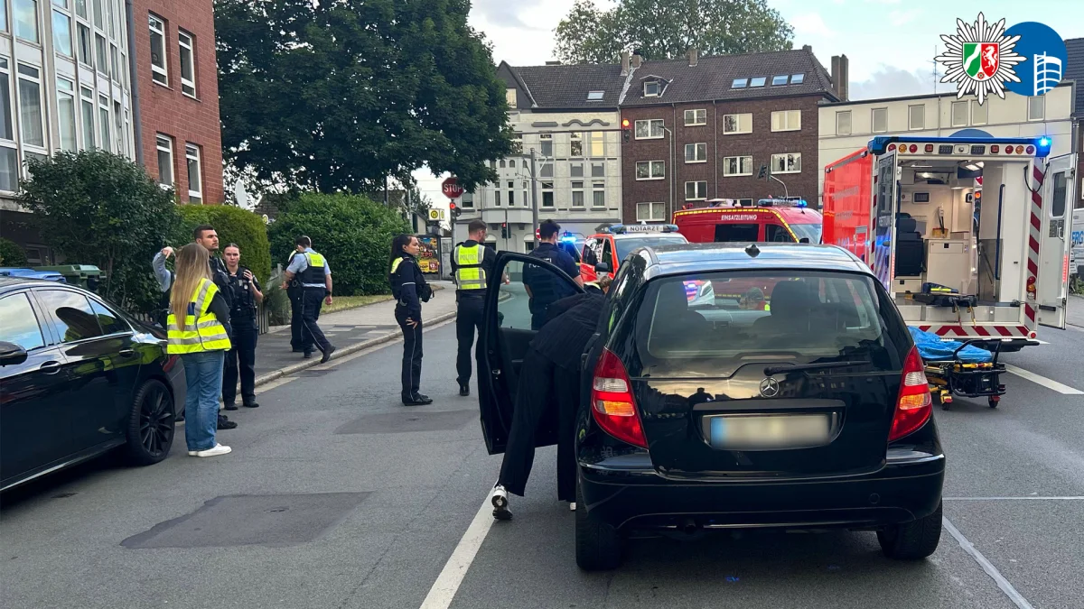 Mehrere Polizisten und ein Rettungsfahrzeug sind auf einer Straße im Einsatz. Im Vordergrund steht ein schwarzer Mercedes.