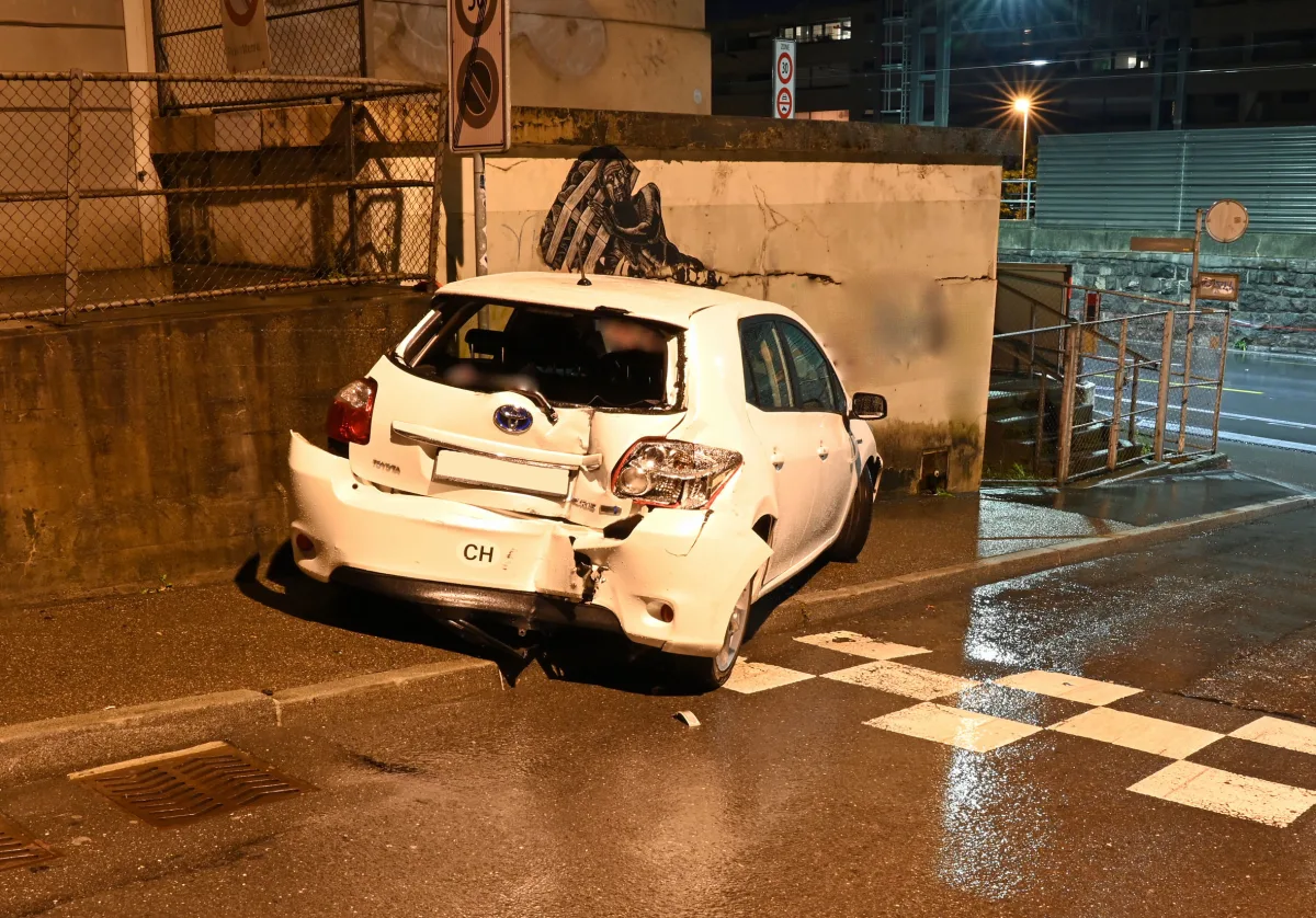 Ein weißer Kleinwagen mit deutlichem Heckschaden steht bei Nacht vor einer Mauer. Die Straße ist nass.