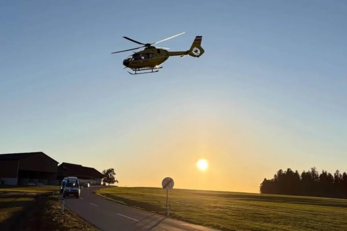 Ein Rettungshubschrauber fliegt bei Sonnenuntergang über eine ländliche Straße, inmitten einer offenen Landschaft mit Feldern und einem Waldrand im Hintergrund.