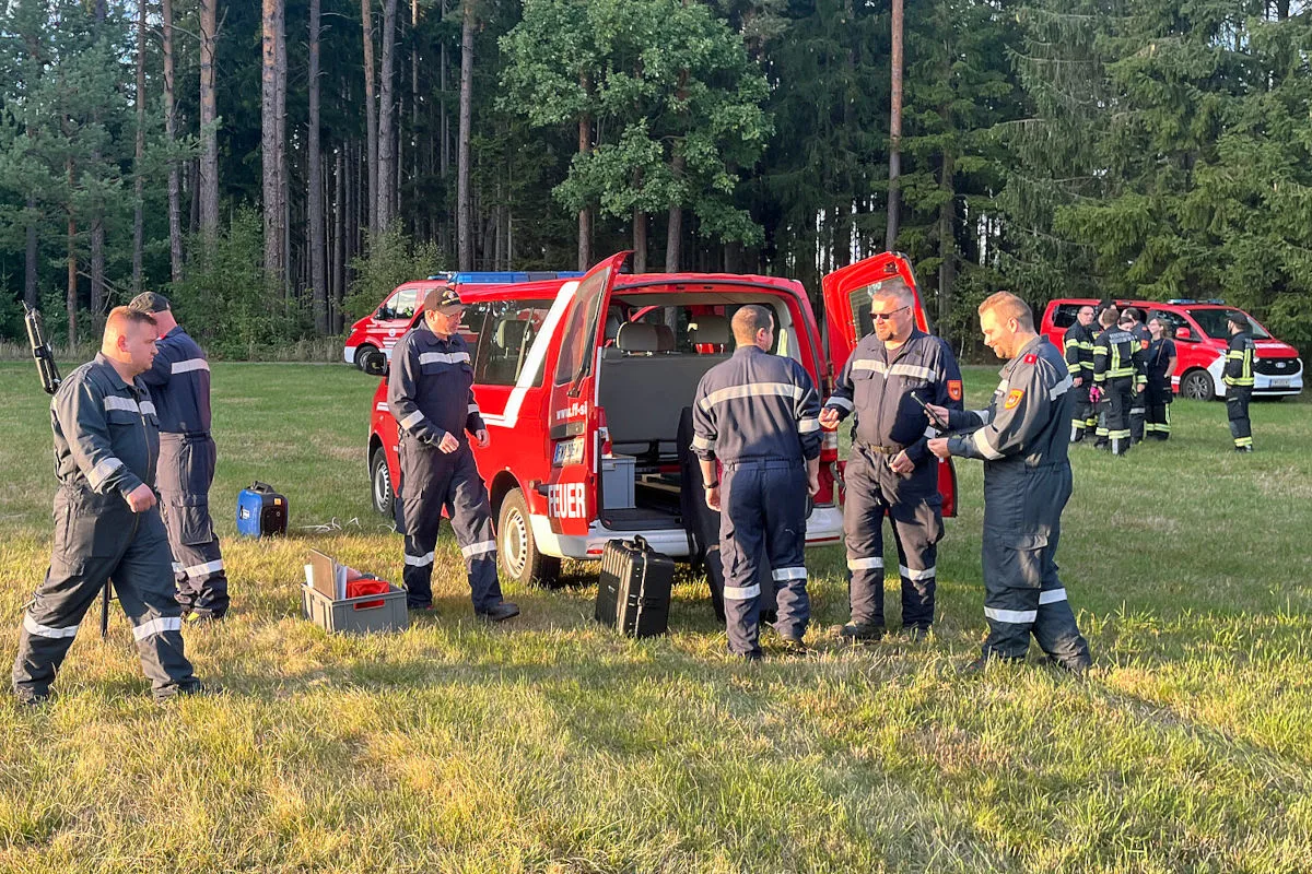 Mehrere Feuerwehrleute stehen neben einem roten Einsatzfahrzeug mit offenem Kofferraum auf einer Wiese vor einem Wald. Weitere Einsatzkräfte und Fahrzeuge sind im Hintergrund zu sehen.