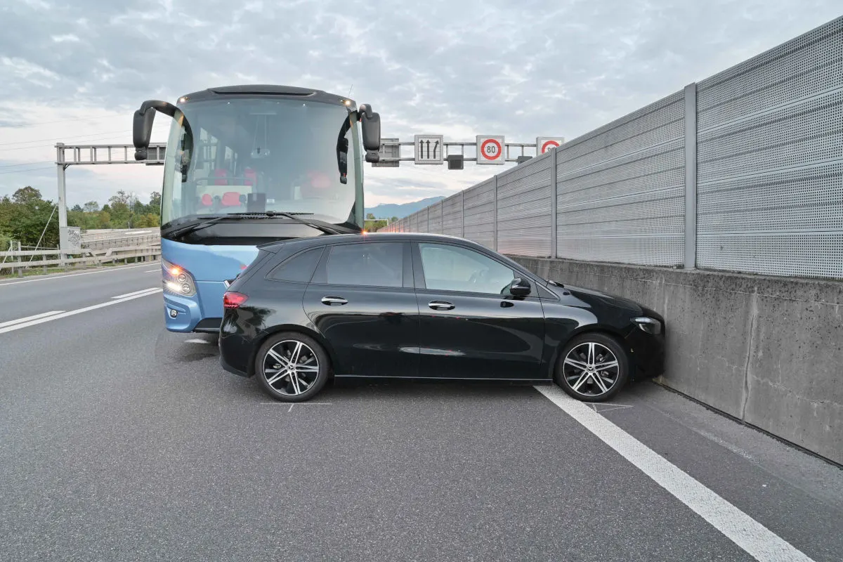 Ein schwarzes Auto steht quer vor einem blauen Linienbus auf einer Autobahn und ist an eine Leitplanke gedrückt.