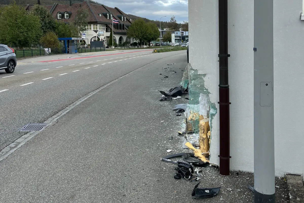 Eine Gebäudeecke ist stark beschädigt und von mehreren Autoteilen umgeben, die auf dem Gehweg verstreut liegen.