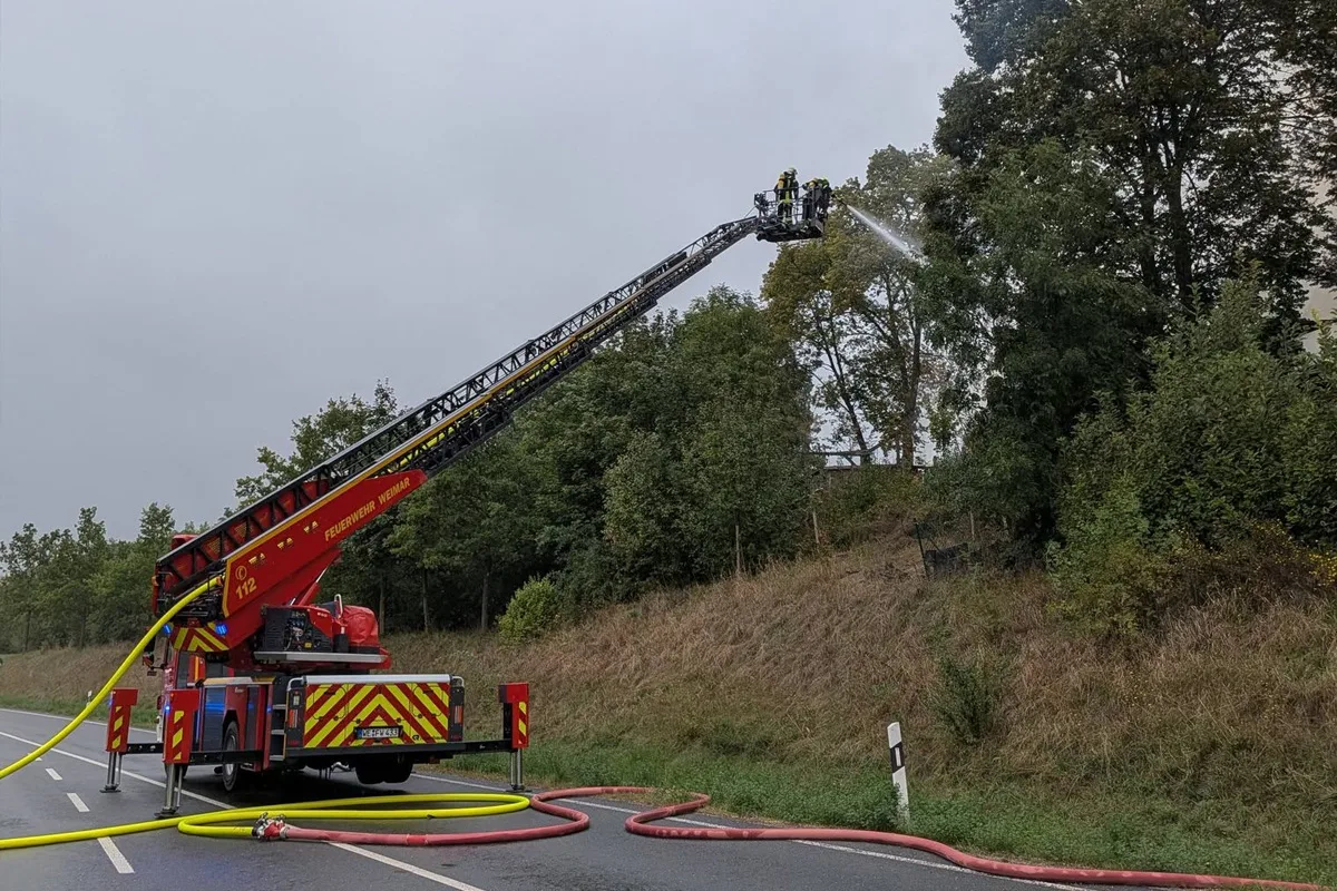 Ein Feuerwehrfahrzeug mit der Aufschrift 'Feuerwehr Weimar' steht am Straßenrand, während Feuerwehrmänner auf einer Drehleiter Wasser auf einen brennenden Baum sprühen.