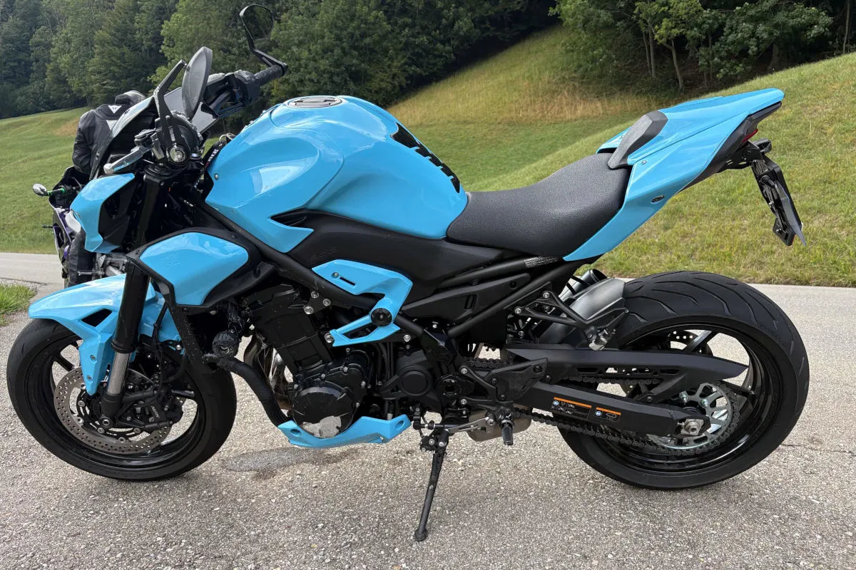 Ein blaues Motorrad steht auf einem asphaltierten Weg neben einer Grünfläche, rechts sind Bäume zu sehen.