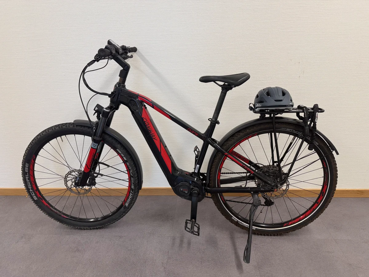 Ein schwarzes E-Bike mit roten Details steht seitlich vor einer weißen Wand. Ein Fahrradhelm liegt auf dem Gepäckträger.