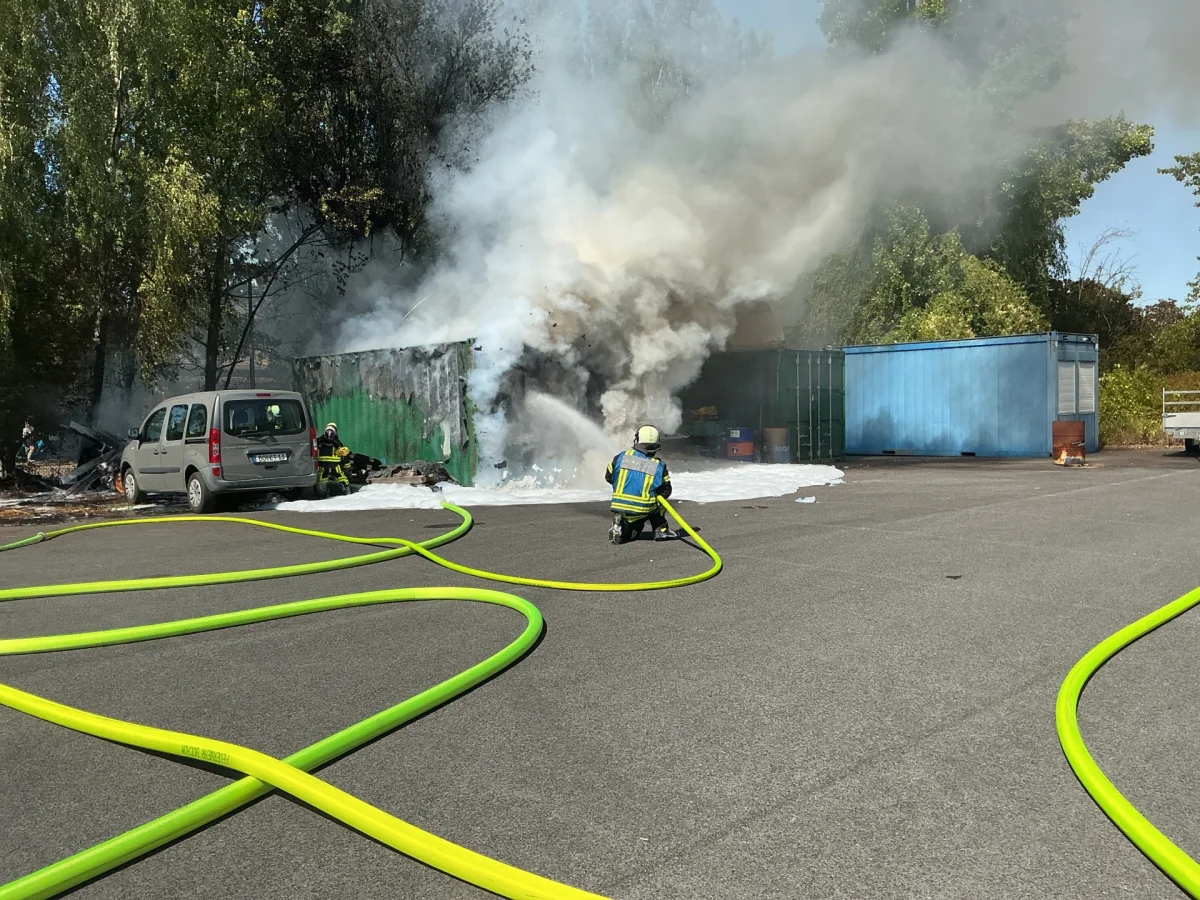 Zwei Feuerwehrleute bekämpfen einen Containerbrand mit Löschschläuchen. Dichter Rauch steigt auf.