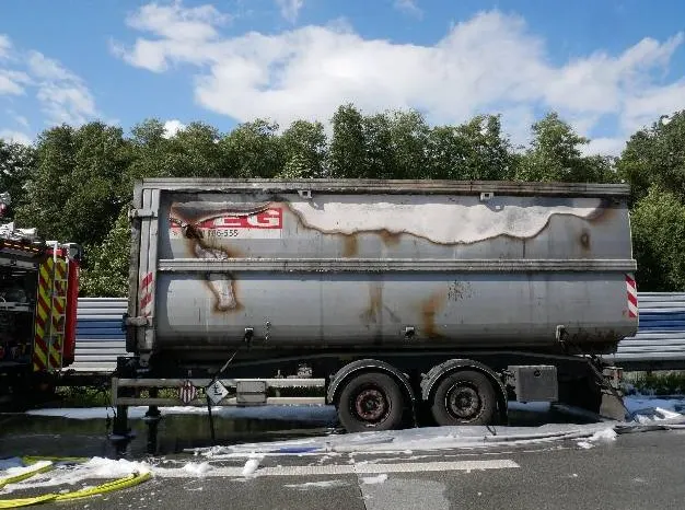 Ein Lastwagenanhänger mit starken Brandspuren steht auf einer Autobahn. Im Hintergrund sind Bäume und ein blauer Himmel zu sehen.