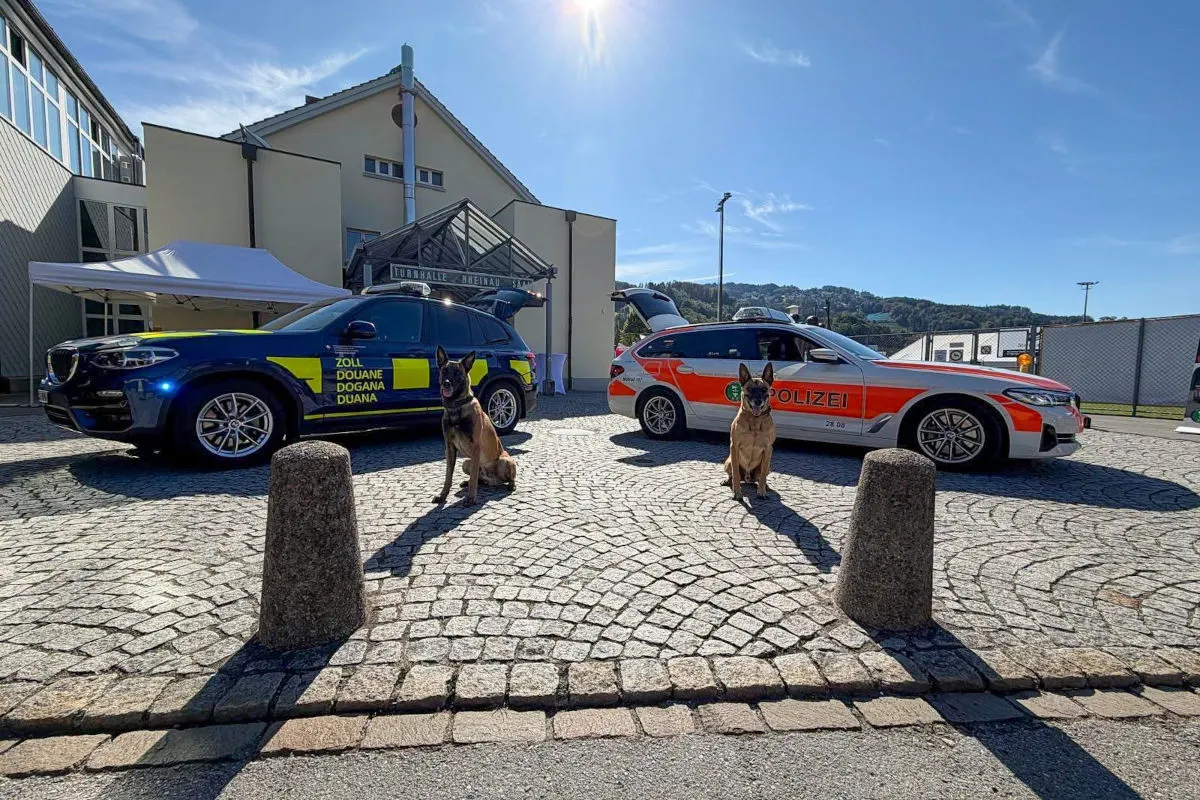 Ein Zollfahrzeug zu linken und ein Polizeifahrzeug zu rechten, beide mit Hunden, vor einem modernen Gebäude bei Sonnenschein.