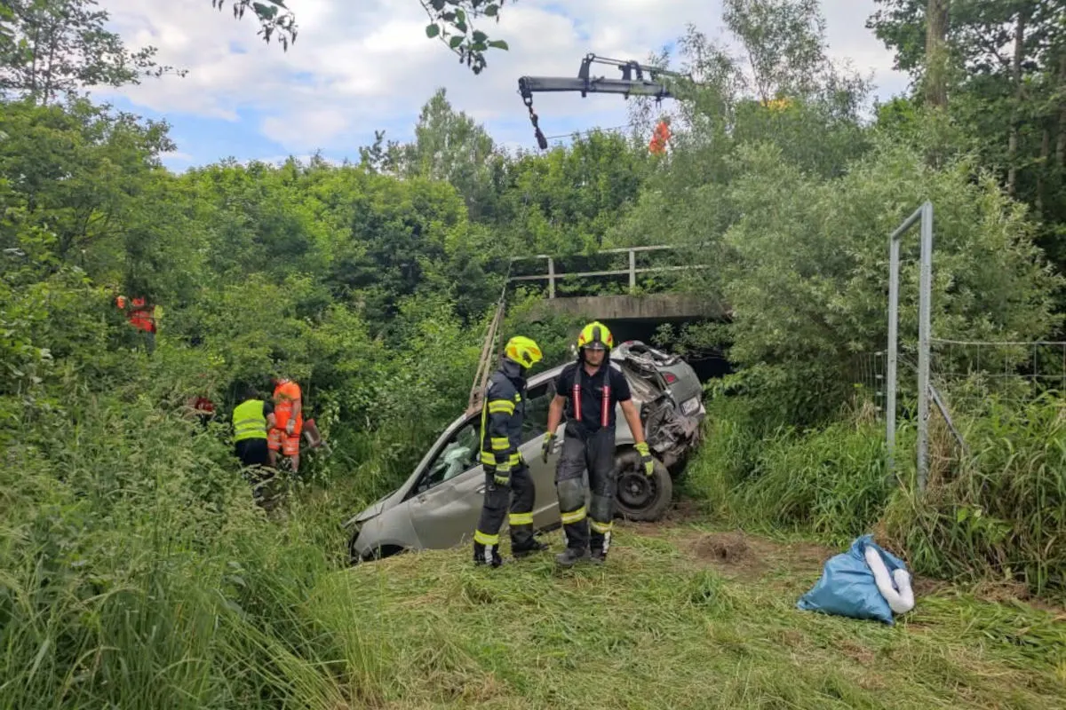 Ein Auto liegt verunfallt in einem bewachsenen Graben. Feuerwehrleute stehen vor dem Fahrzeug und führen Bergearbeiten durch. Eine Brücke ist im Hintergrund sichtbar.