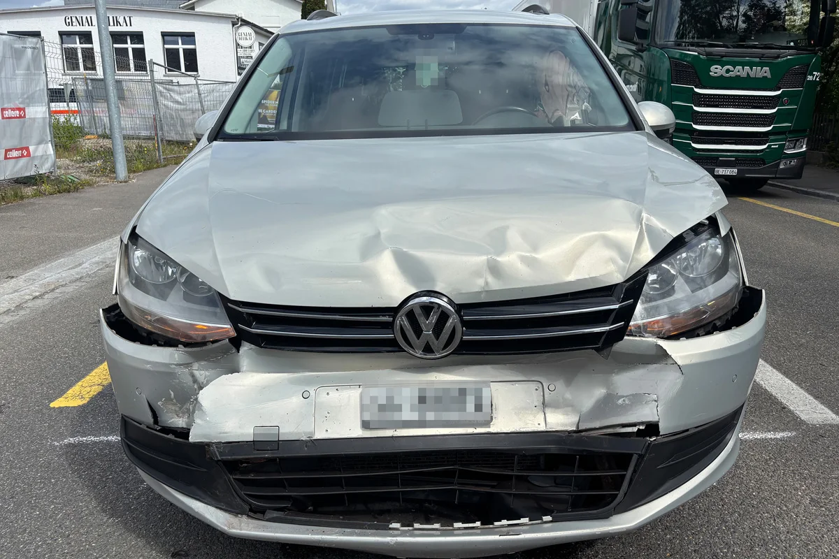 Ein silberner VW mit erheblichen Beschädigungen an der Front steht auf einer Straße. Im Hintergrund ist ein grüner Lastwagen teilweise sichtbar.