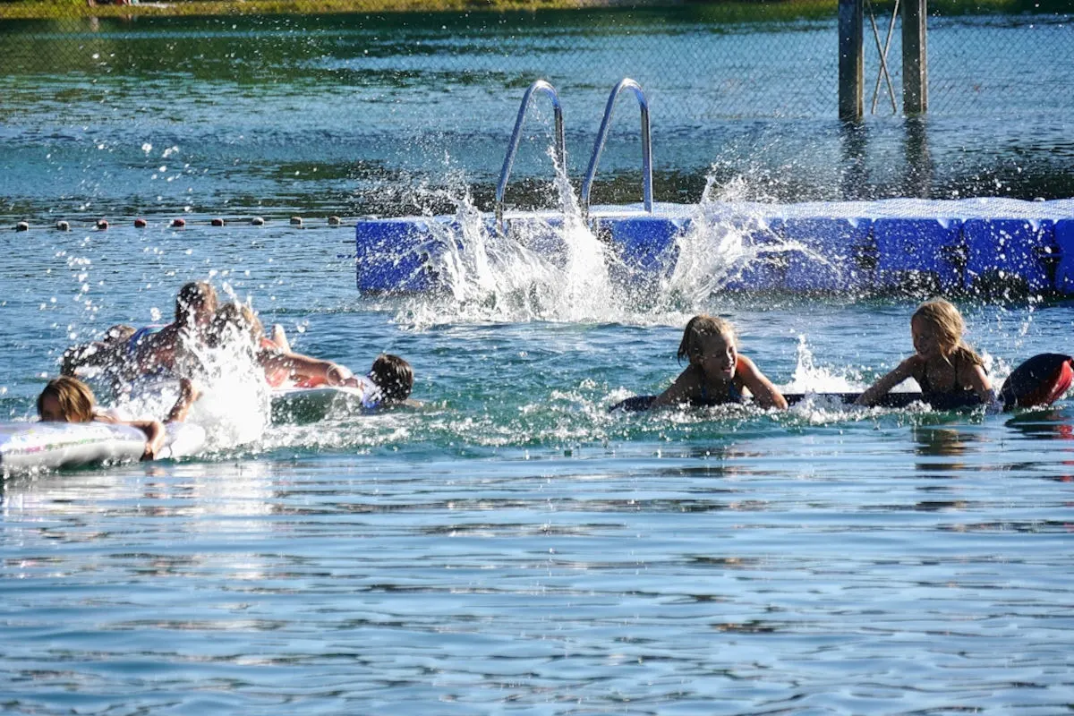 Mehrere Kinder sind in einem Freibad zu sehen, während sie auf dem Wasser planschen. Der blaue Himmel und die Sonne deuten auf einen warmen Tag hin.
