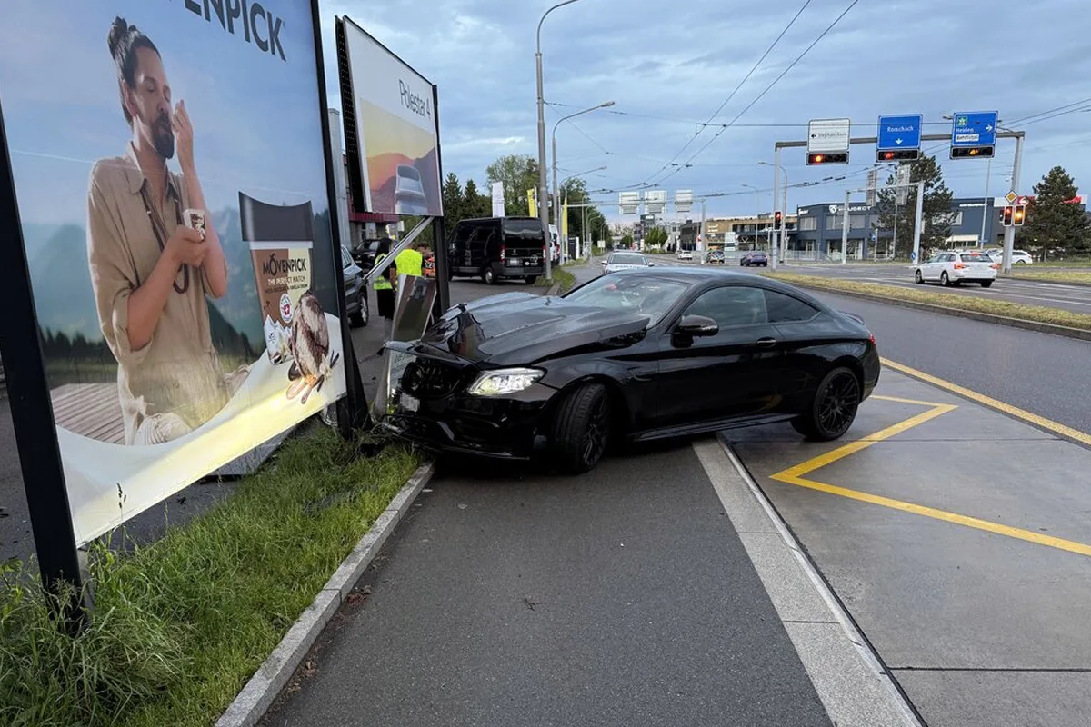 Ein schwarzes Auto hat eine Werbetafel gerammt und steht mit beschädigter Front an einer Stadtkreuzung. Ein weiteres Fahrzeug ist im Hintergrund sichtbar.