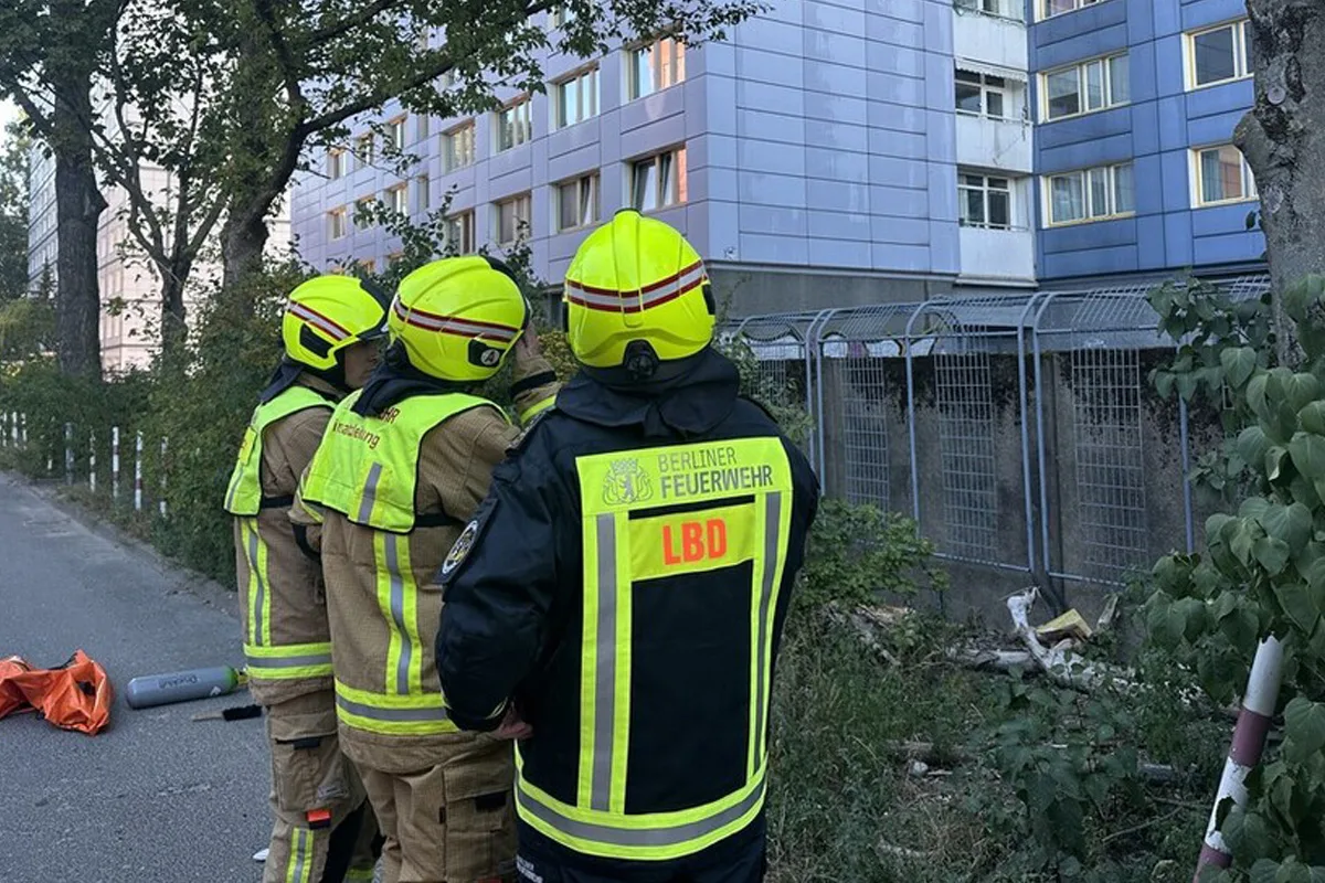 Drei Feuerwehrleute in Schutzkleidung stehen vor einem hohen Wohngebäude. Sie tragen Helme und reflektierende Jacken mit der Aufschrift Berliner Feuerwehr.