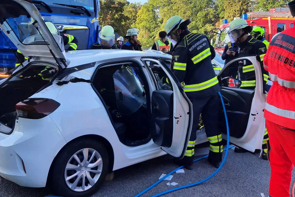 Feuerwehrleute der Berufsfeuerwehr München sind dabei, ein beschädigtes, weißes Auto nach einem Unfall zu sichern. Rettungskräfte sind ebenfalls vor Ort.