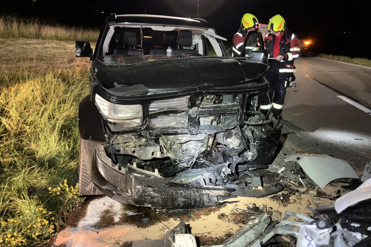 Ein schwarzes Auto mit stark beschädigter Frontpartie steht neben einer Straße. Im Hintergrund stehen Feuerwehrleute in reflektierenden Anzügen.