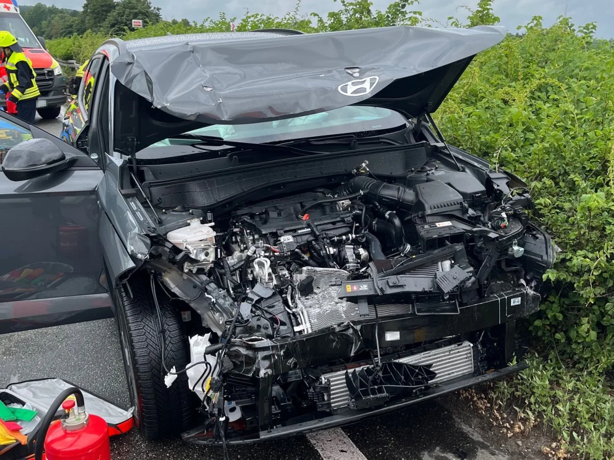 Ein Hyundai mit zerstörter Front steht am Straßenrand. Ein Rettungsfahrzeug und Personen in Uniform sind im Hintergrund sichtbar.
