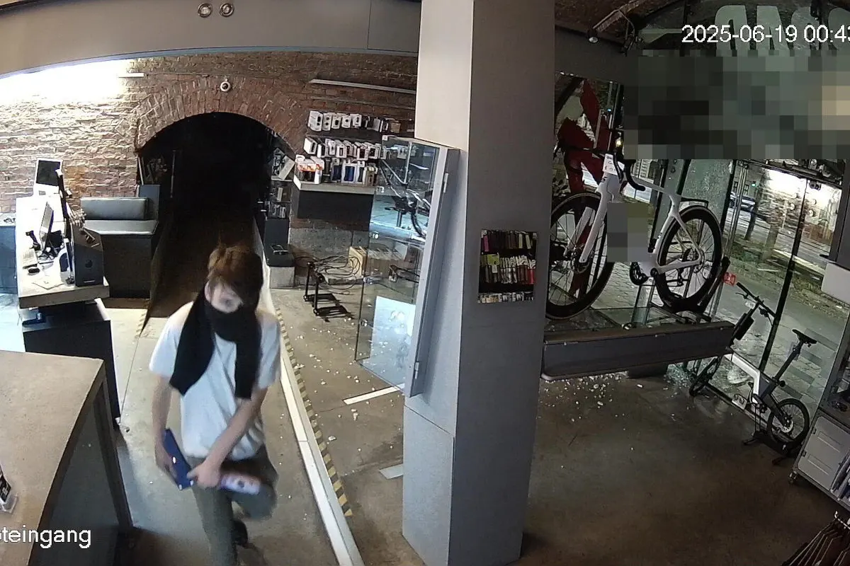 Ein maskierter Mensch hastet durch den Innenraum eines Fahrradgeschäfts mit gestohlenen Waren. Im Hintergrund sind Schaufenster und Ausstellungsräder zu sehen.