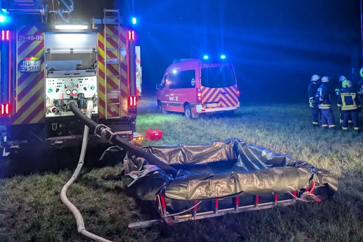 Zwei Feuerwehrautos stehen auf einem Feld im Einsatz, verbunden durch Wasserschläuche. Feuerwehrleute sind anwesend.
