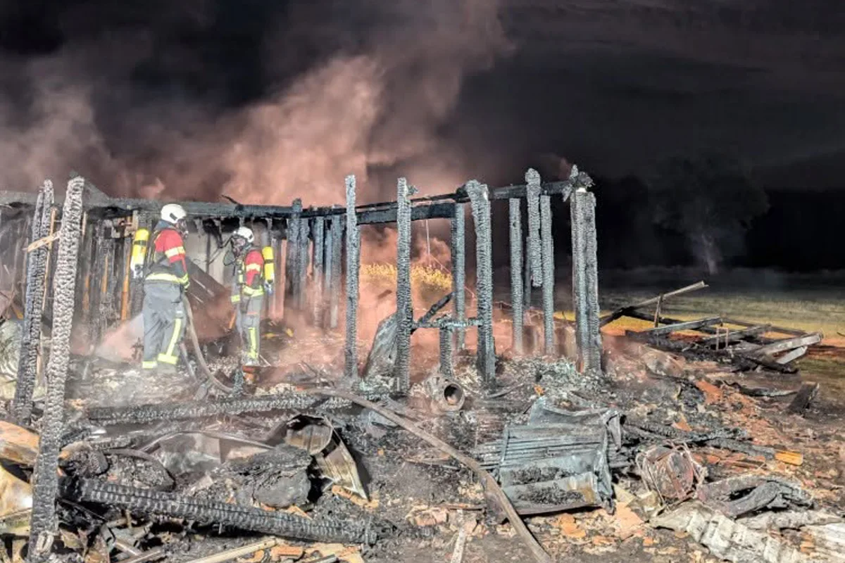 Zwei Feuerwehrleute in Schutzkleidung arbeiten in einem brennenden, stark beschädigten Holzgebäude. Rauch steigt auf.