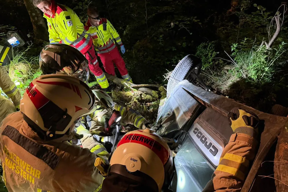 Mehrere Feuerwehrleute und ein Rettungssanitäter sind bei Nacht um ein umgestürztes Fahrzeug im Wald versammelt. Das Auto liegt auf der Seite und Rettungsmaßnahmen sind im Gange.