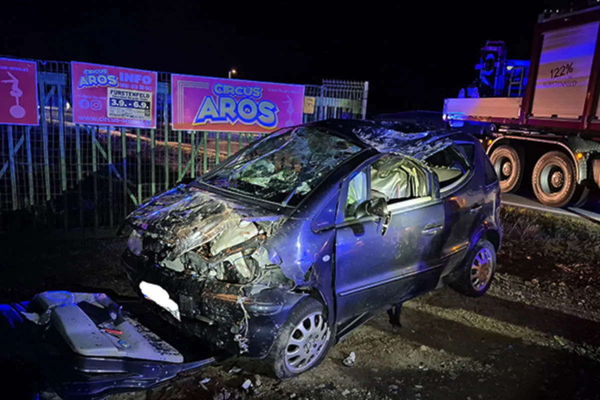 Ein stark beschädigter Pkw steht neben einem Lkw auf einem Zirkusgelände bei Nacht. Der Wagen hat deutliche Schäden an Front und Dach.