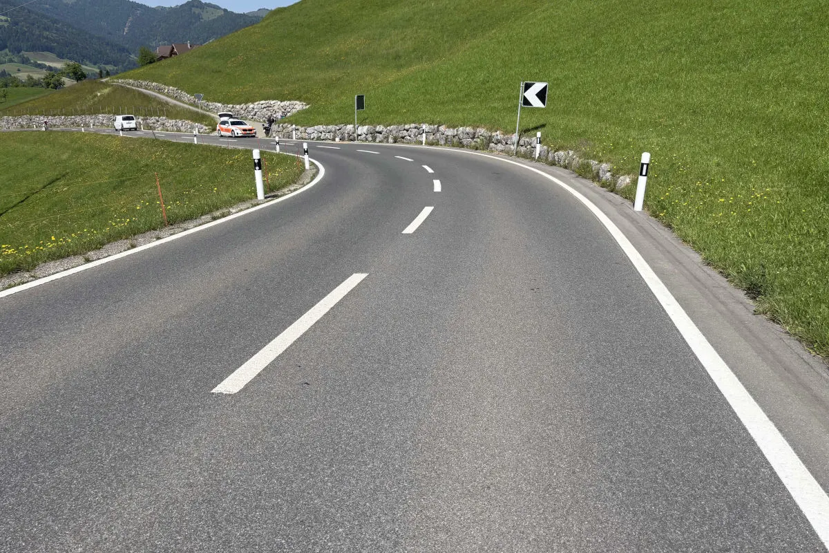 Auf einer kurvigen Landstraße sind ein Polizeiauto und mehrere andere Fahrzeuge zu sehen. Die Straße führt durch eine grüne, hügelige Landschaft bei klarem Wetter.
