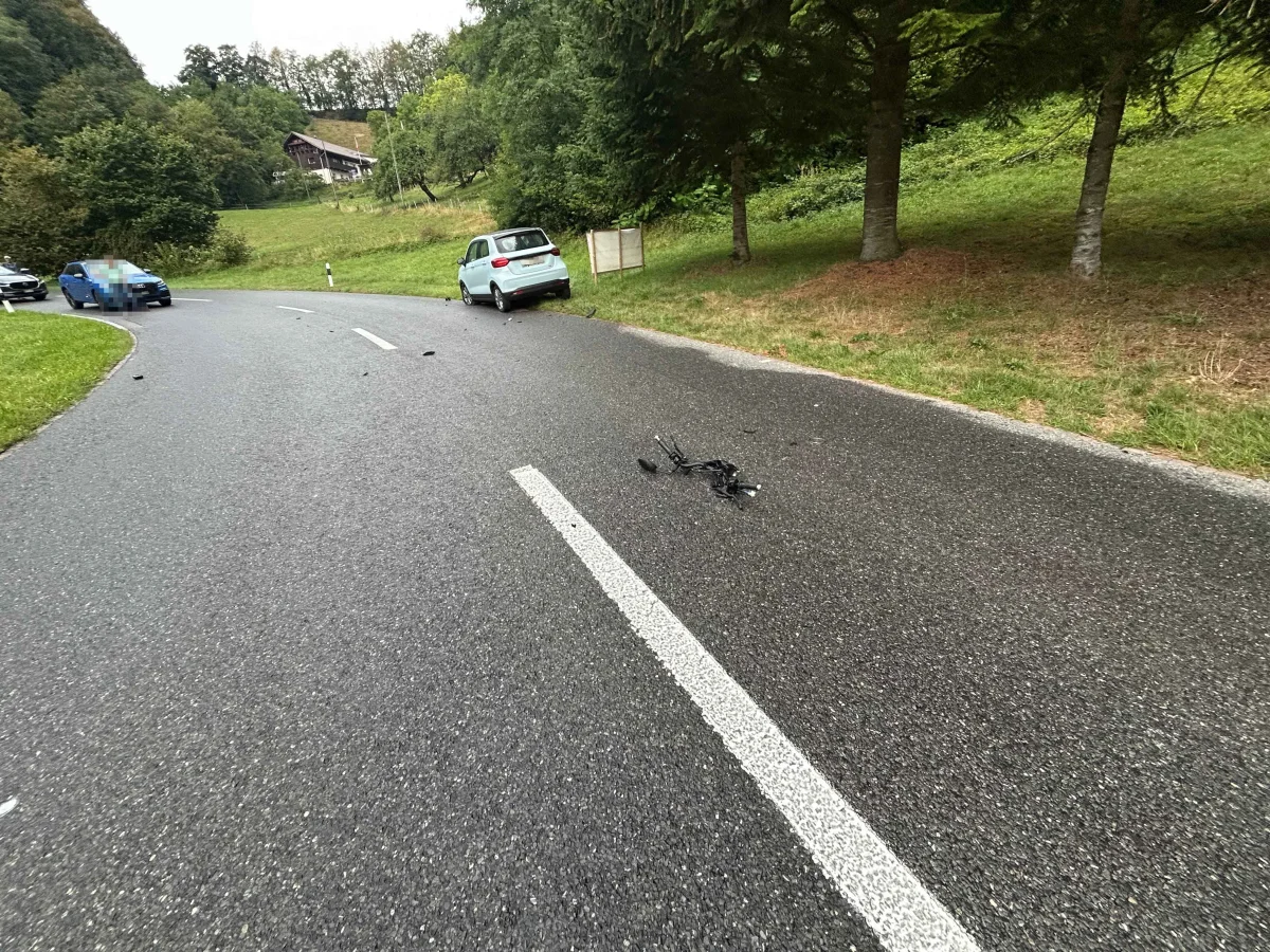 Auf einer kurvigen Landstraße liegen Trümmer. Ein Auto steht am Straßenrand; Bäume und ein Gebäude im Hintergrund.