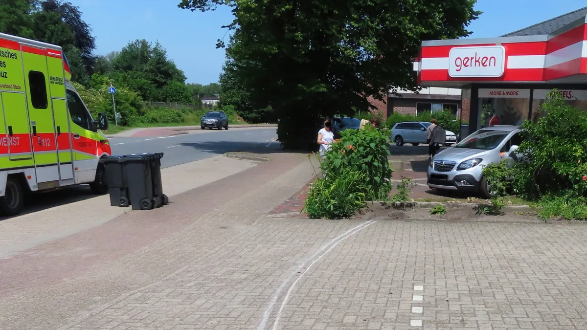 Ein gelb-roter Rettungswagen steht am Rand einer Straße vor einem Gebäude mit der Aufschrift 'gerken'. Vor dem Gebäude stehen zwei Frauen auf dem Gehweg.