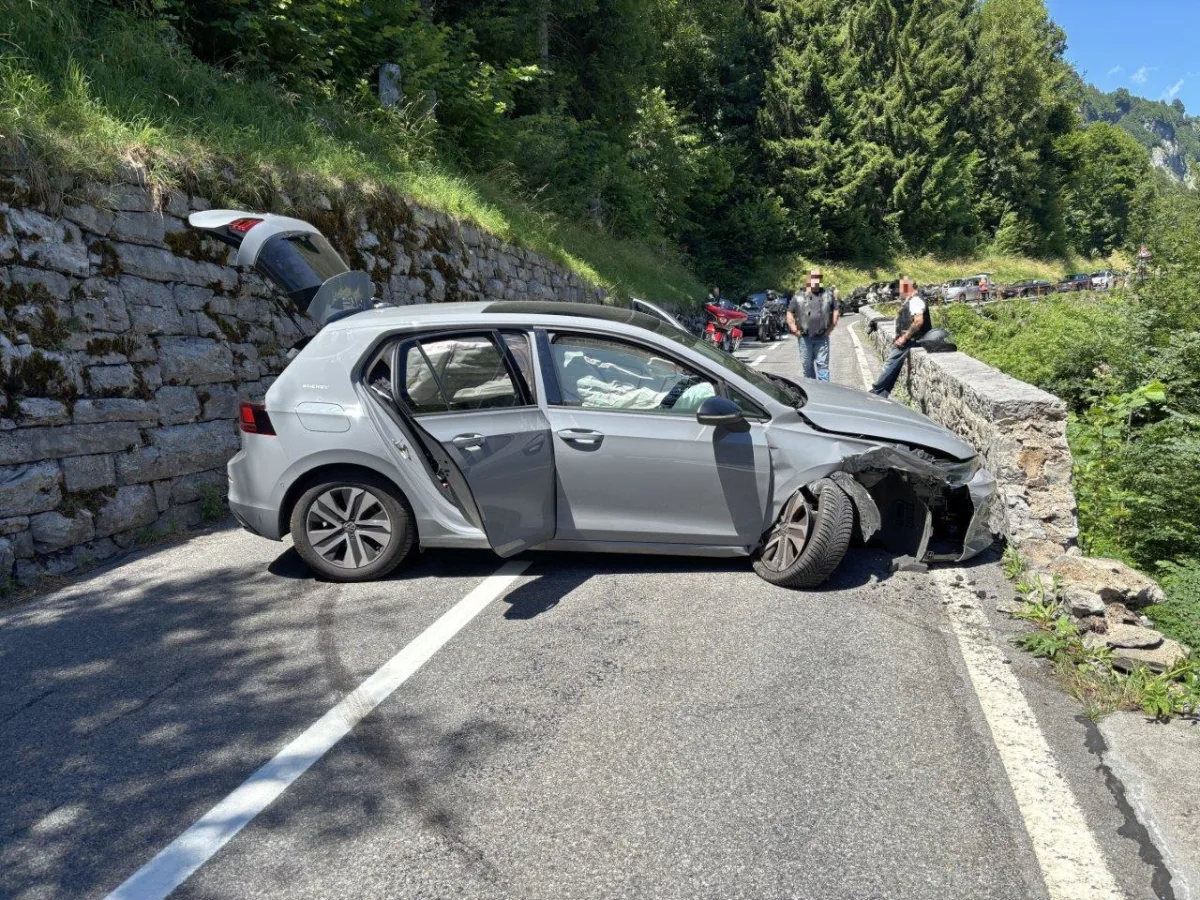Ein graues Auto mit geöffneter Fahrertür steht nach einer Kollision mit einer Steinmauer auf einer Landstraße. Personen stehen in der Nähe der Unfallstelle.