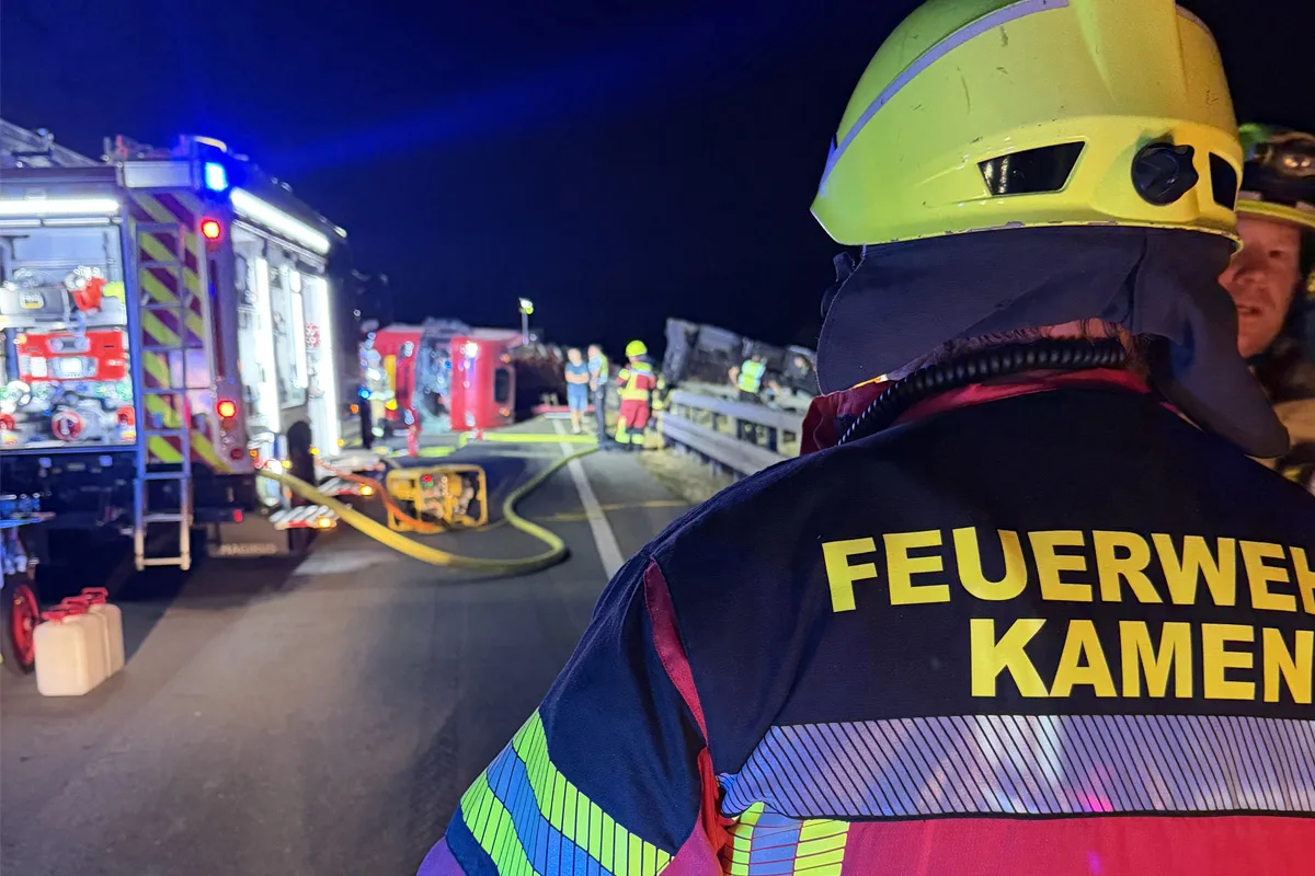 Ein Feuerwehrmann steht im Vordergrund in gelber Ausrüstung. Im Hintergrund ist ein umgestürztes Fahrzeug und ein weiteres Feuerwehrfahrzeug mit eingeschaltetem Blaulicht zu sehen.