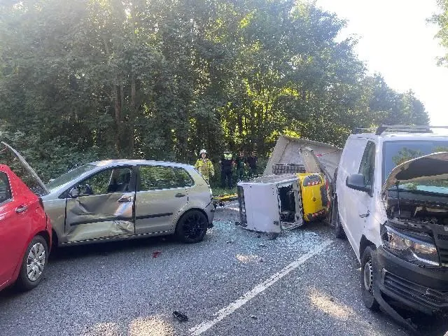 Drei Fahrzeuge sind bei einem Unfall auf einer Landstraße beschädigt. Im Hintergrund stehen Bäume und Einsatzpersonal in gelber Kleidung.