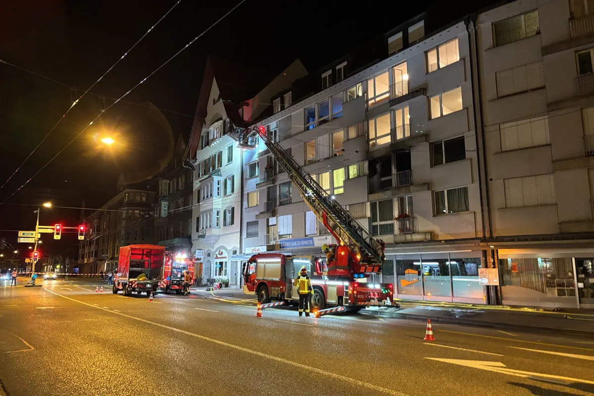 Ein Feuerwehrauto mit Drehleiter ist vor einem mehrfach Stockwerken hohen Wohngebäude im Einsatz, um einen Brand zu bekämpfen. Die Straße ist abgesperrt und Feuerwehrautos sind zu sehen.