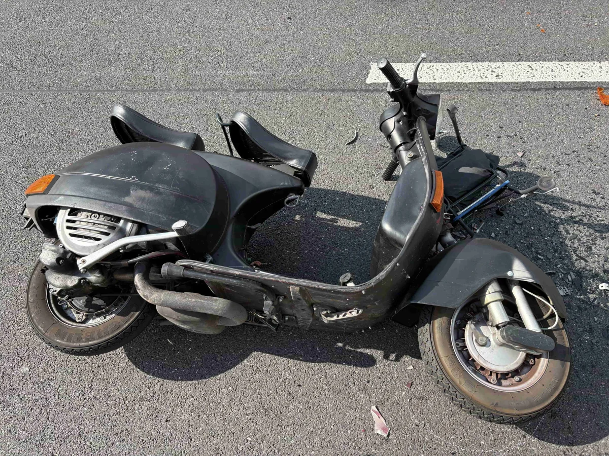 Auf dem Bild ist ein schwarzer Motorroller zu sehen, der auf der Straße umgestürzt ist. Teile des Rollers, darunter der Lenker und die Verkleidung, sind beschädigt.