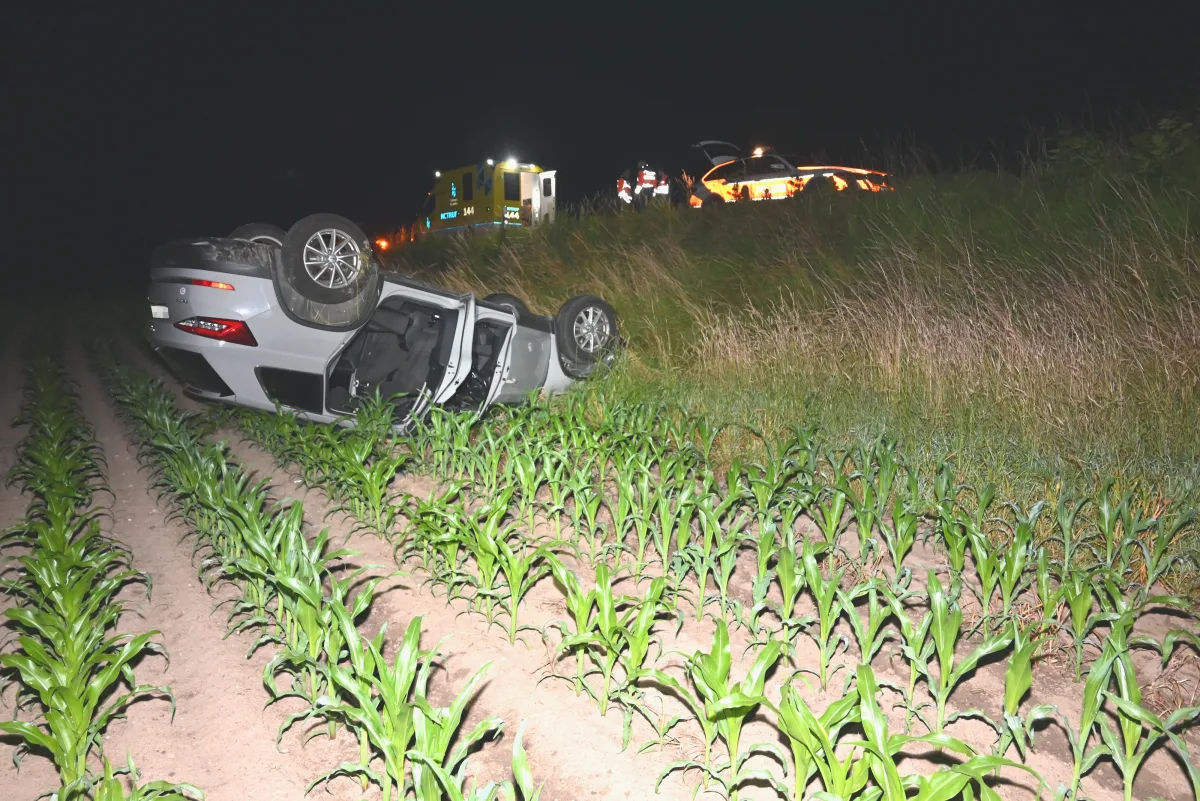 Bei Nacht ist ein Auto in einem Feld auf dem Dach liegend sichtbar. Im Hintergrund stehen Rettungskräfte mit einem Fahrzeug.