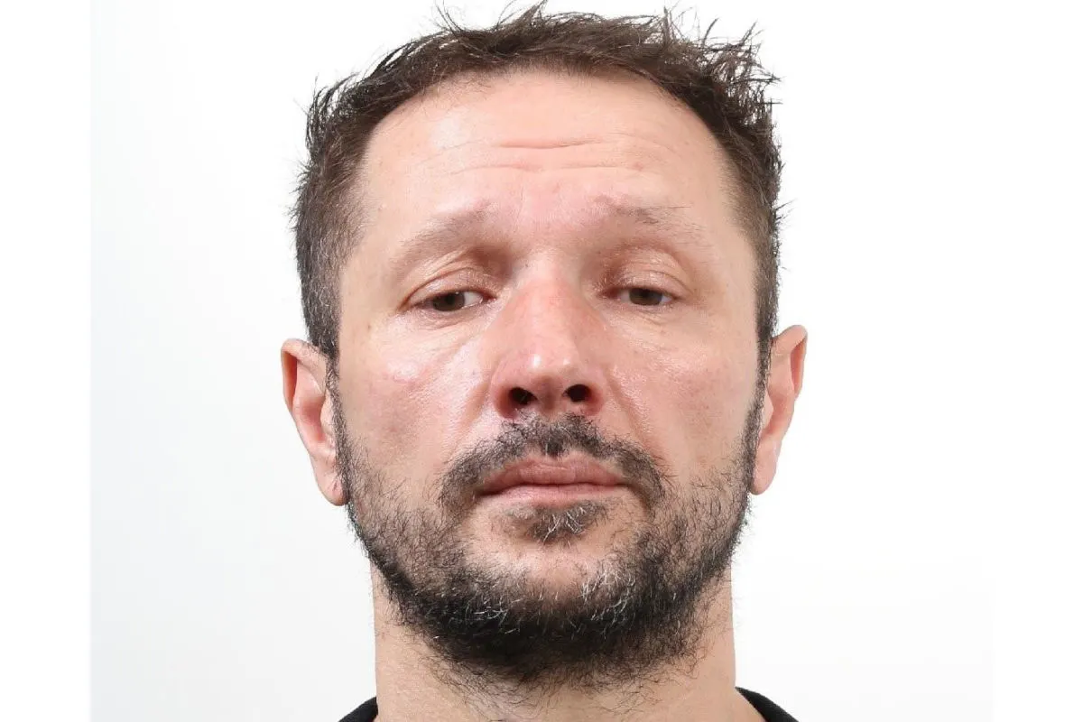 Das Bild zeigt das Gesicht eines Mannes mit kurzem dunklen Haar und Bart vor einem weißen Hintergrund. Es handelt sich um ein Porträtfoto, vermutlich im Fahndungskontext.