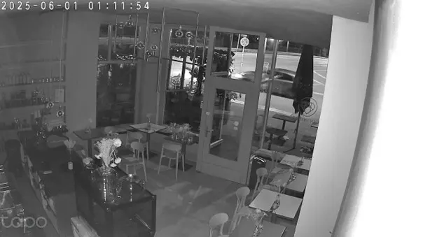 Das Bild einer Überwachungskamera zeigt ein menschenleeres Restaurant und ein Auto, das nachts draußen vorbeifährt. Die Szene ist in Schwarz-Weiß gehalten und spiegelt ruhige Nachtverhältnisse wider.