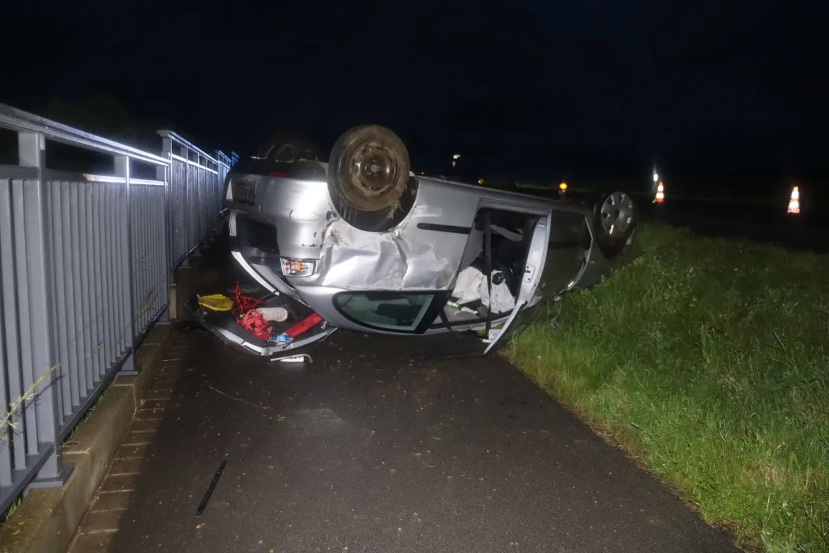 Ein silbernes Auto liegt auf dem Dach auf einer nassen Straße. Verkehrshütchen sind im Hintergrund zu sehen.