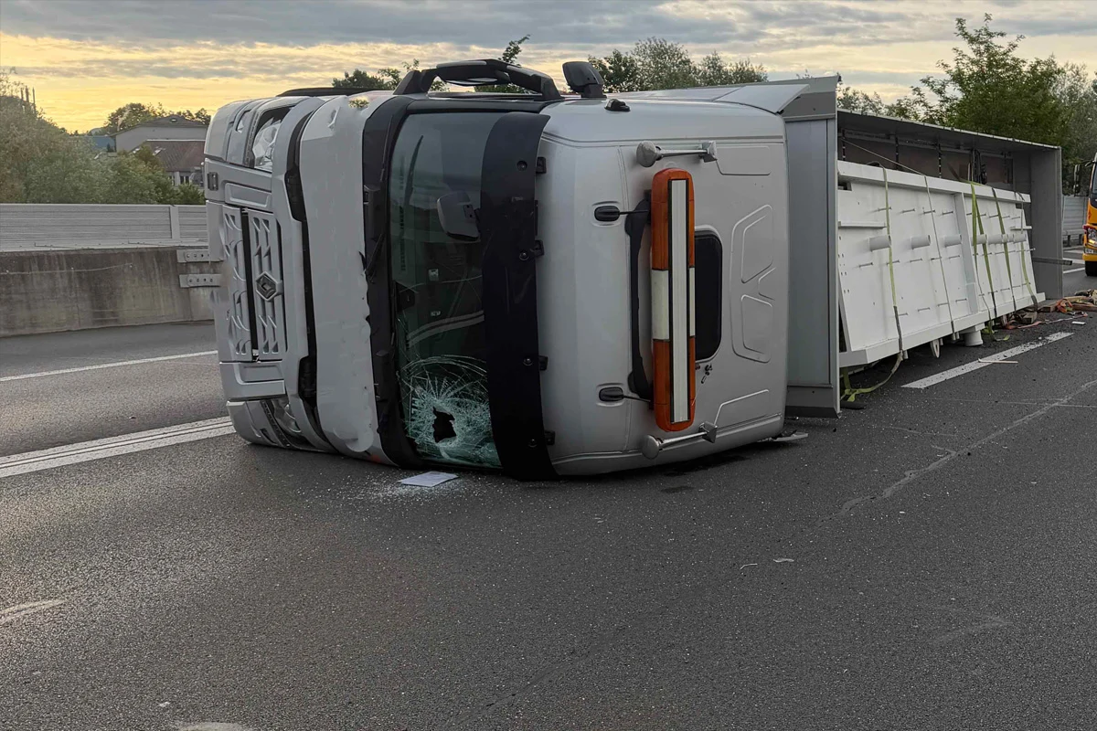 Ein Lkw liegt auf der Seite auf der Autobahn und blockiert eine Fahrspur. Die Frontscheibe ist beschädigt, und es gibt keine sichtbaren Personen.