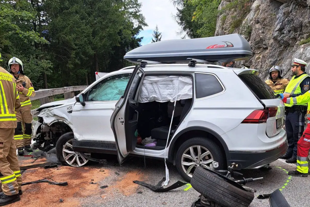 Ein weißes Auto mit sichtbaren Schäden steht auf der Straße, umgeben von Feuerwehrleuten und Polizisten. Der Airbag ist ausgelöst und Trümmer liegen auf dem Boden.