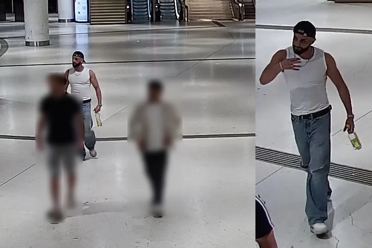 Der Mann trägt ein weißes T-Shirt und Jeans, hält eine Flasche und befindet sich in einer geräumigen Halle mit Treppen im Hintergrund.